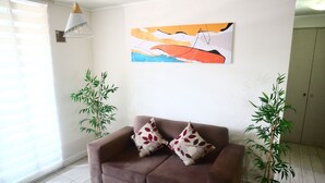 A living room with a brown sofa, two decorative pillows, a painting on the wall, and two potted plants. - Tranquilo Departamento Full Equipado (Santiago)