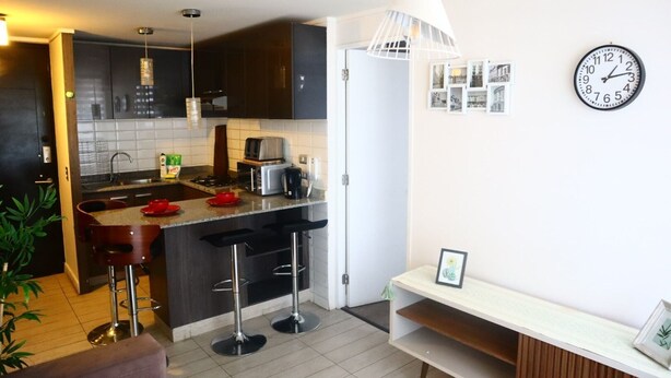 A modern kitchen with dark cabinets, a central island, bar stools, a clock, and a framed picture on the wall. - Tranquilo Departamento Full Equipado (Santiago)