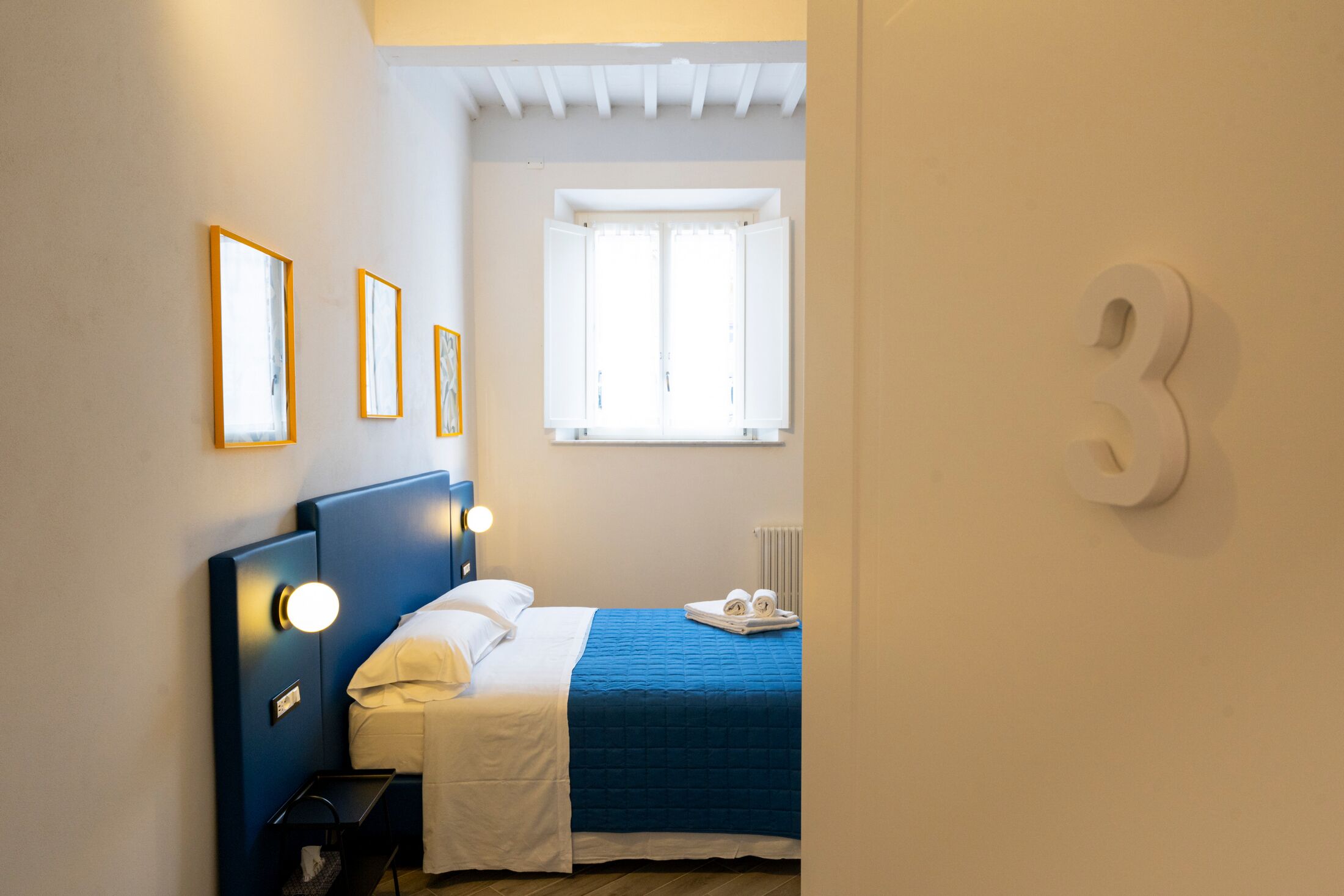 Quarto com uma cama, mesas de cabeceira, janela e um número fixado na parede.