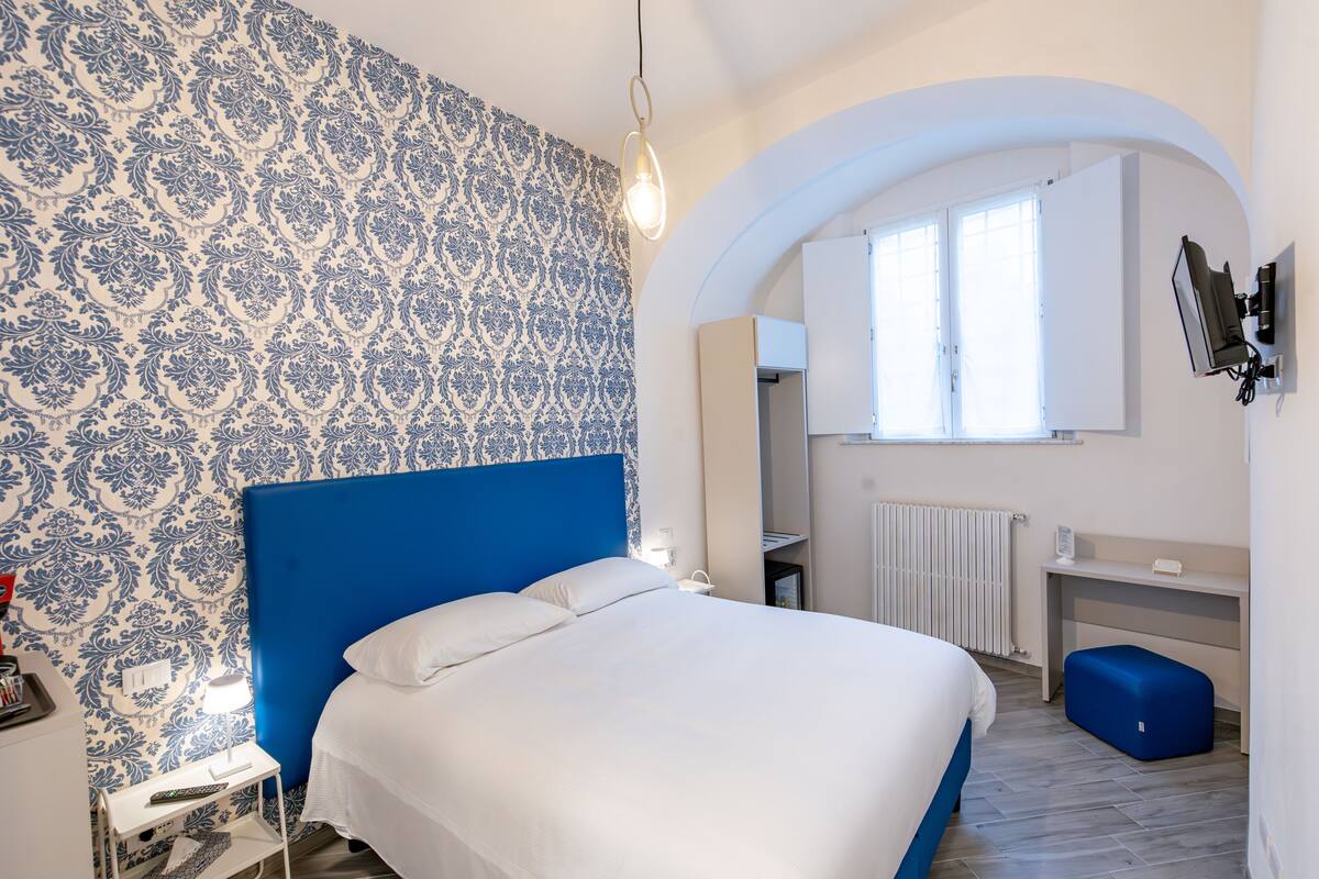 Een slaapkamer met een blauw hoofdeinde, wit beddengoed, een bureau met stoel en een grote spiegel.