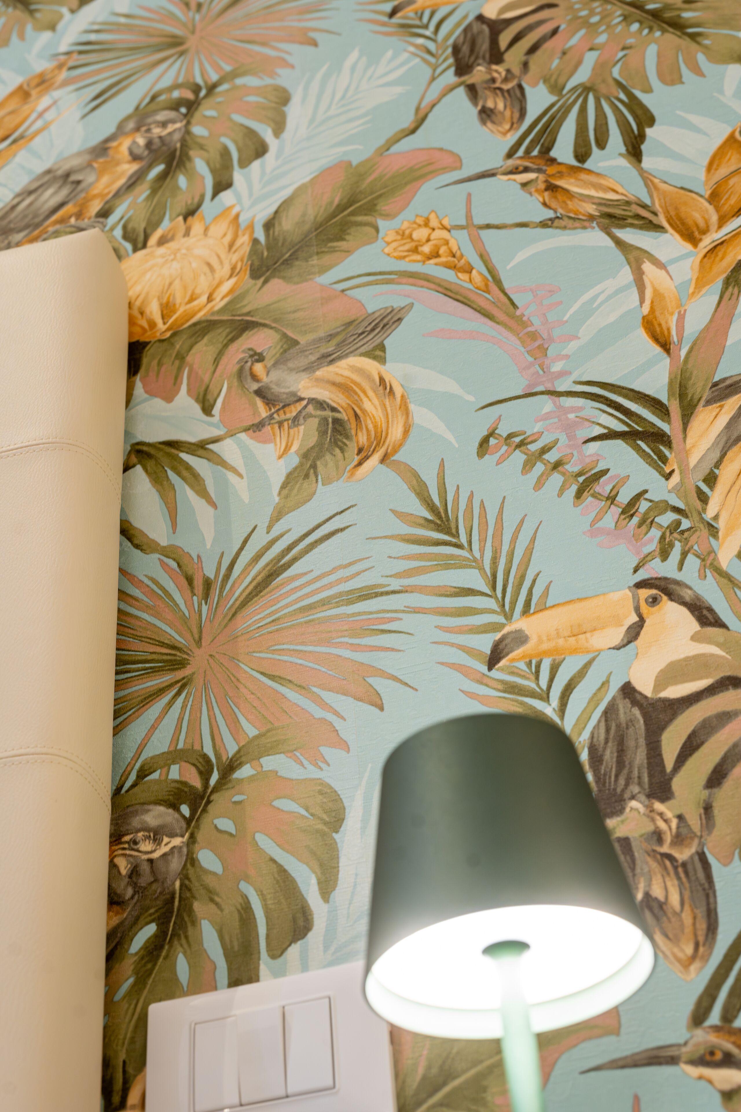 Ein Zimmer mit tropischem Wandpapier, das Vögel und Pflanzen zeigt, einer weißen Schalterverteilung und einer grünen Lampe.