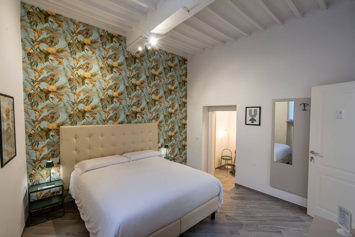 Een slaapkamer met een groot bed, bloemenbehang, een nachtkastje, een lamp en een deur naar een andere kamer.