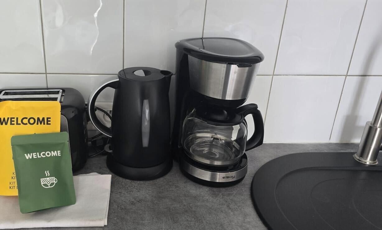 Cozinha com cafeteira, chaleira e torradeira. Há sacolas de boas-vindas no balcão.