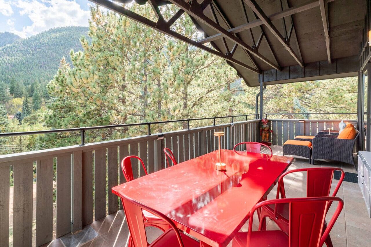 A covered outdoor area with a red table and chairs, a sofa with orange cushions, and a view of a forested mountain range.