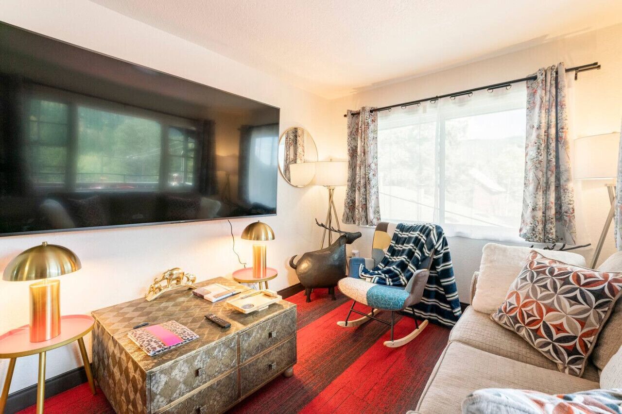 Un salon comprenant un grand téléviseur à écran plat, un canapé, un fauteuil à motifs, une petite table d’appoint avec une lampe et une statuette de cochon décorative.