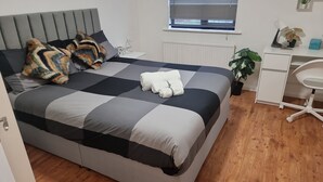 Un lit bien fait, avec une couvre-lit à carreaux, une tête de lit grise et une table de chevet sur laquelle se trouvent une lampe et une plante.