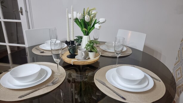 Une table à manger dressée avec de l’assiette, des bols et des couverts blancs, agrémentée d’un vase de fleurs blanches et d’une petite plante en pot.