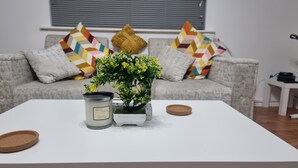 Un salon avec une table basse blanche, une plante en pot, une bougie parfumée à la grenade et un canapé gris agrémenté de coussins colorés.