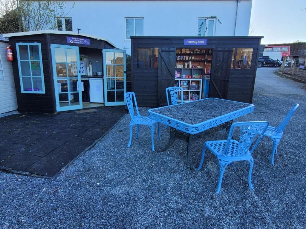 A small outdoor seating area with a table and four chairs, a blue sign, and a building with a sign that reads 'Honesty Shop.'
