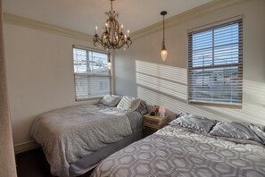 Een slaapkamer met twee bedden, een kroonluchter, een nachtkastje met een lamp en ramen voorzien van zonwering.