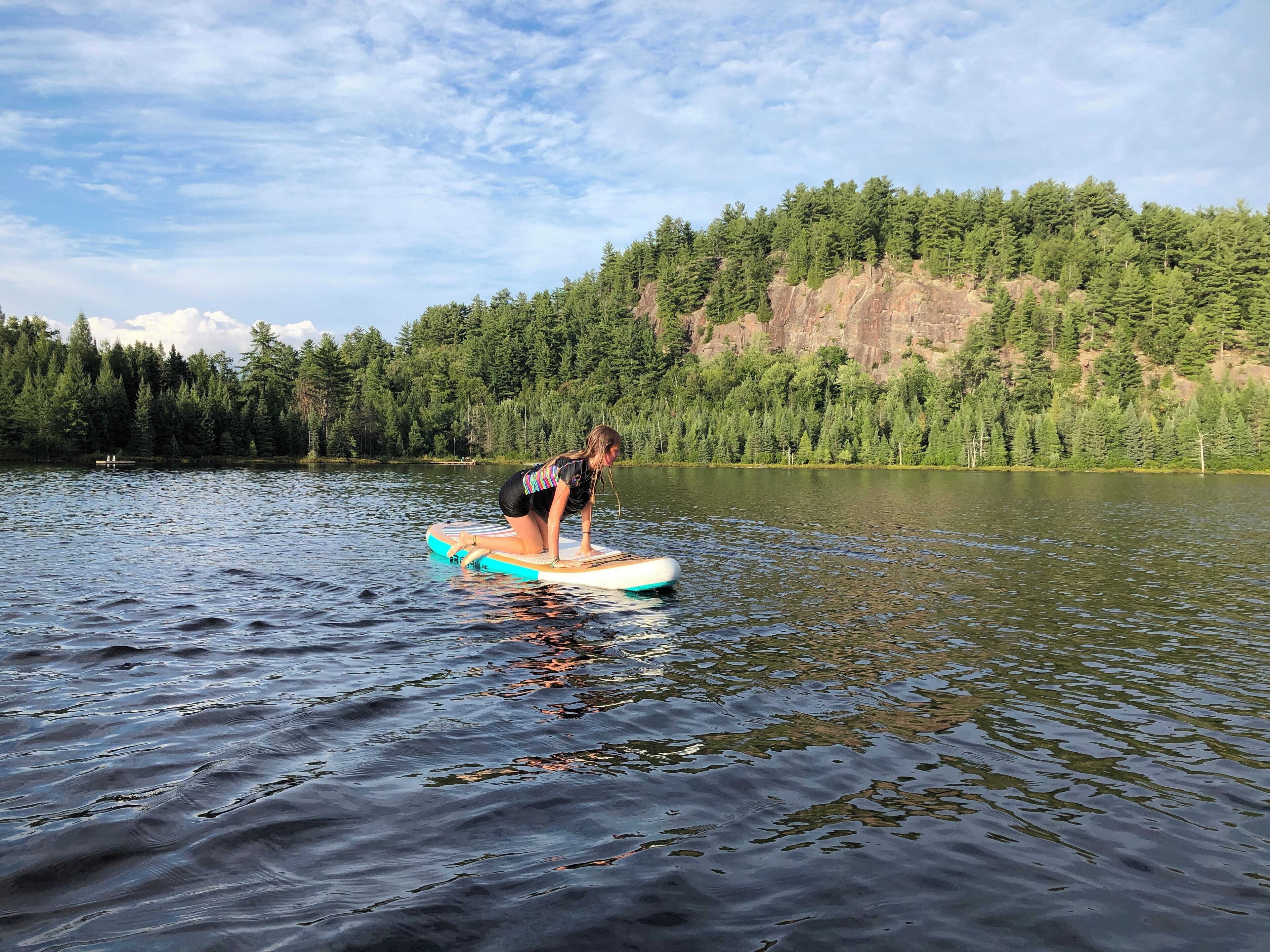 A person on a paddleboard in a calm lake surrounded by dense forest and rocky cliffs.