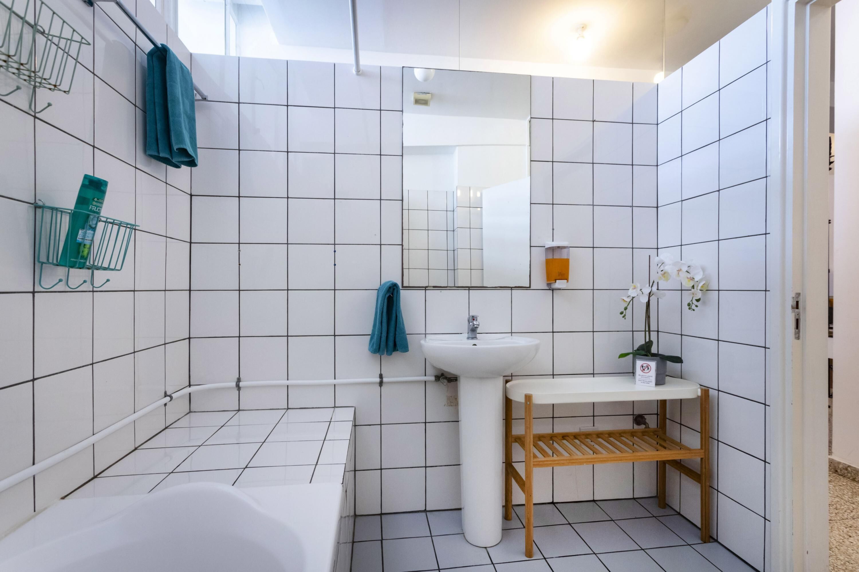 Un baño moderno con azulejos blancos, una bañera, un lavamanos, un espejo y un estante con artículos de aseo.