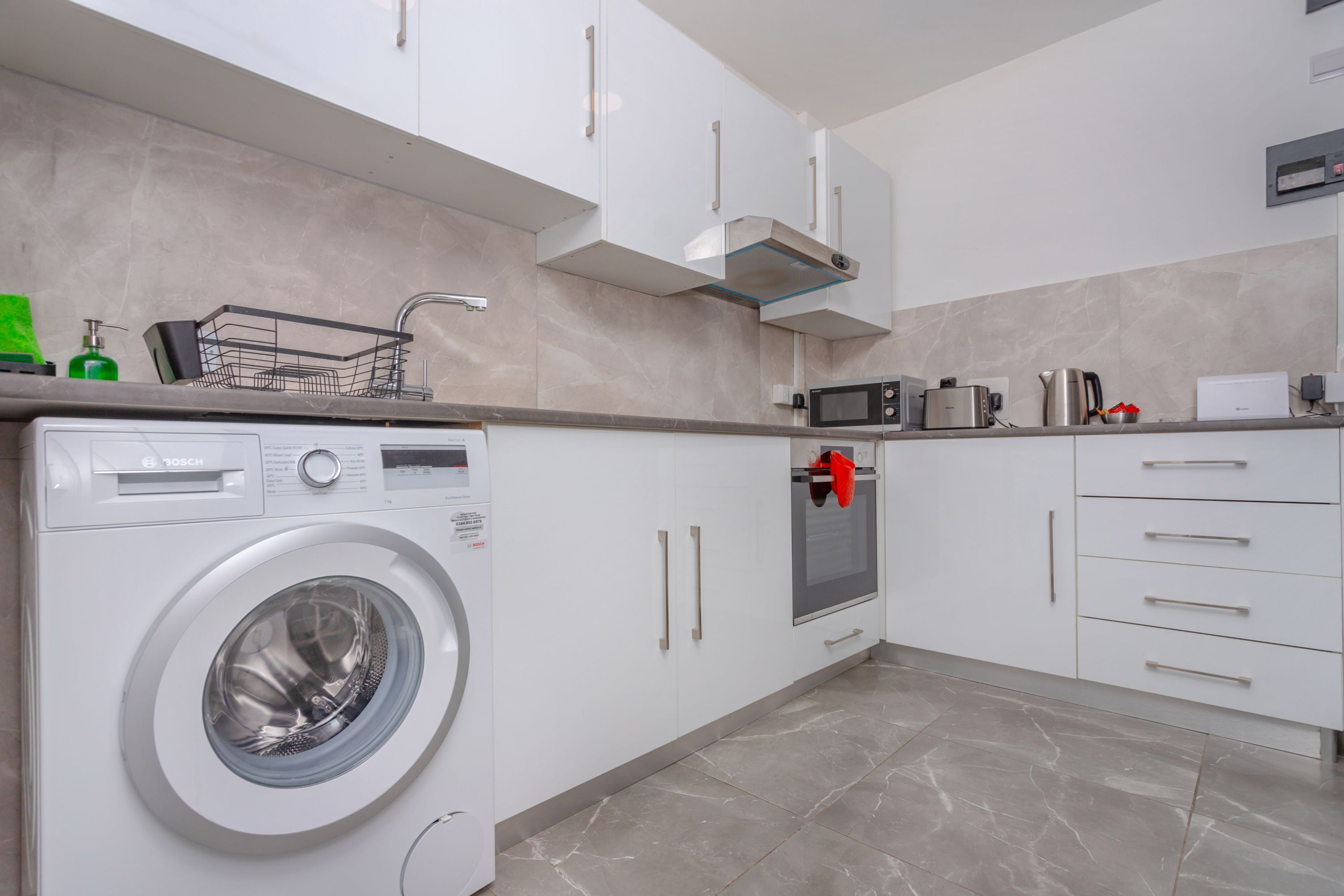 Een moderne keuken met witte kasten, een ingebouwde magnetron, een waterkoker en een wasmachine.
