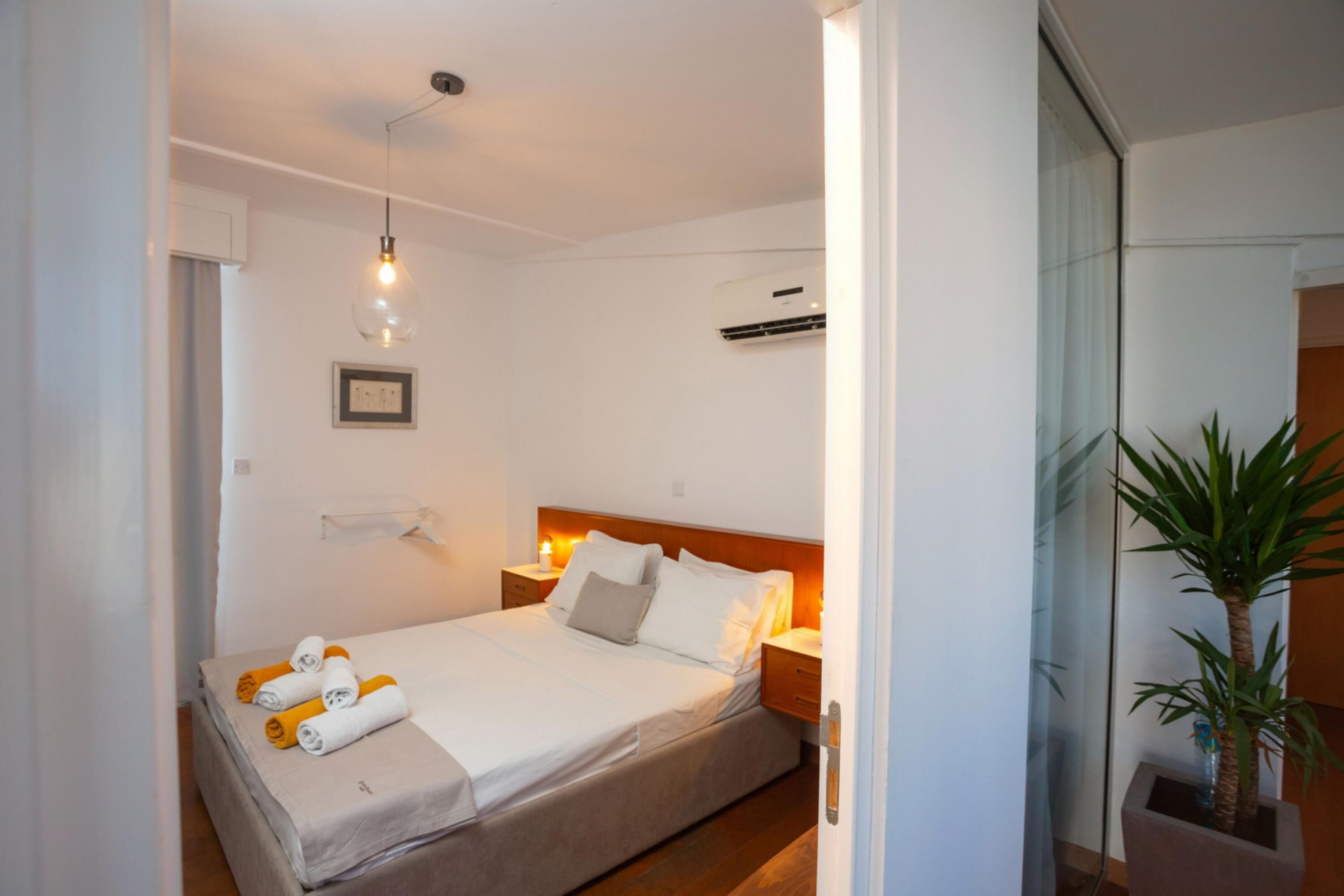 A neatly made bed with towels, a bedside table, a wall-mounted air conditioner, and a potted plant.