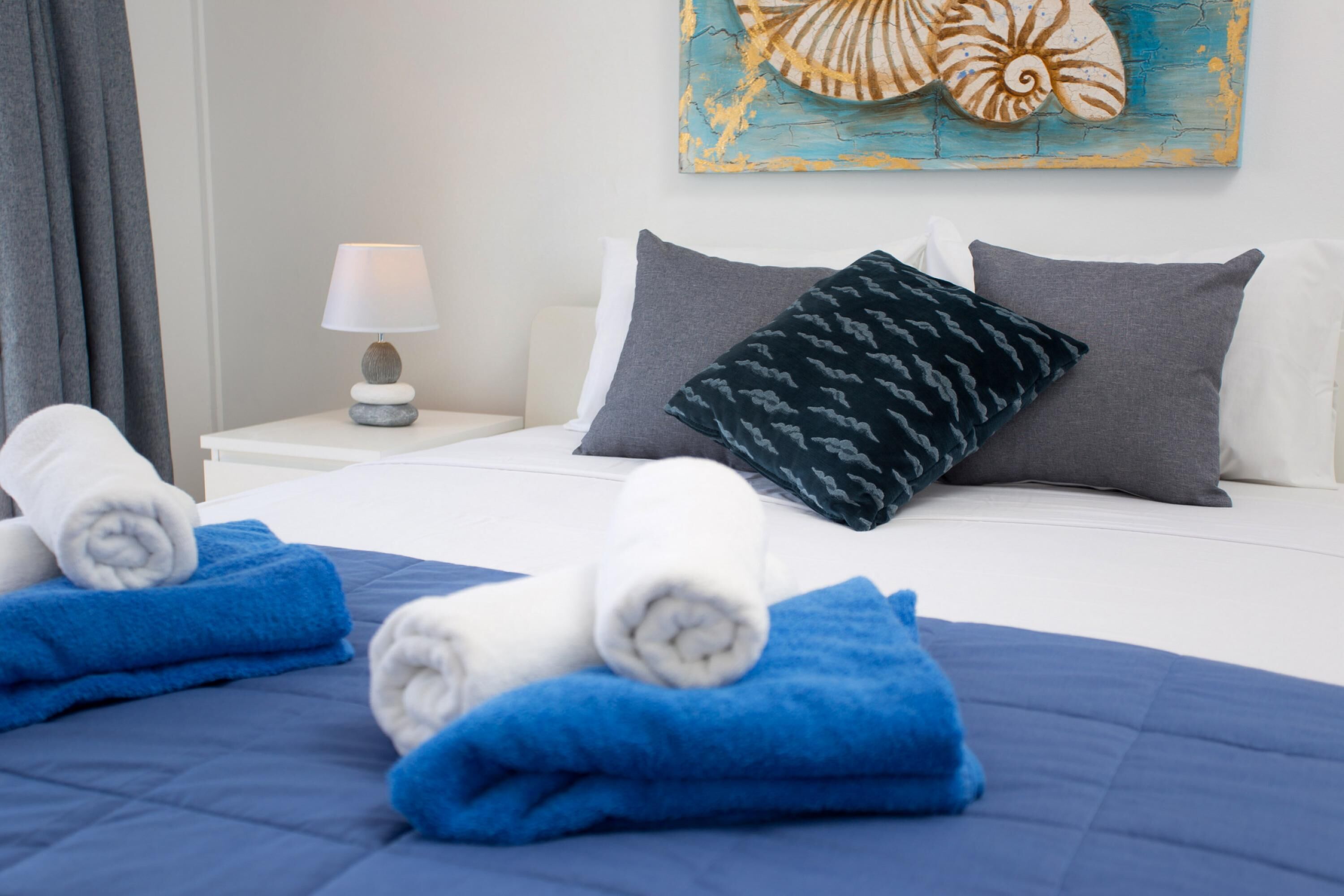 Una cama bien hecha con toallas azules y blancas, una lámpara en la mesita de noche y un cuadro con forma de concha marina en la pared.