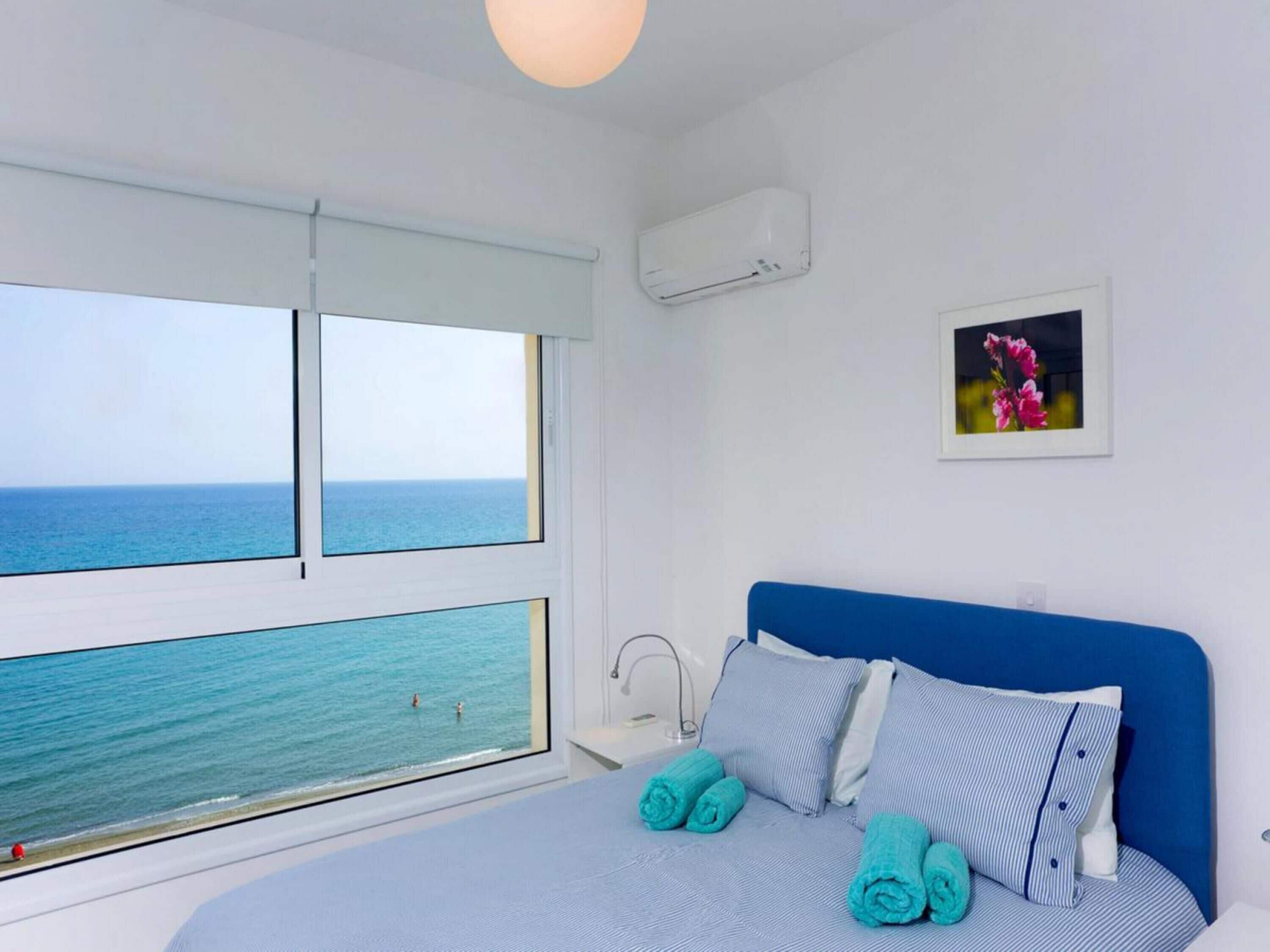 Une chambre avec une grande fenêtre donnant sur la mer, un lit avec une literie bleue et un tableau accroché au mur.