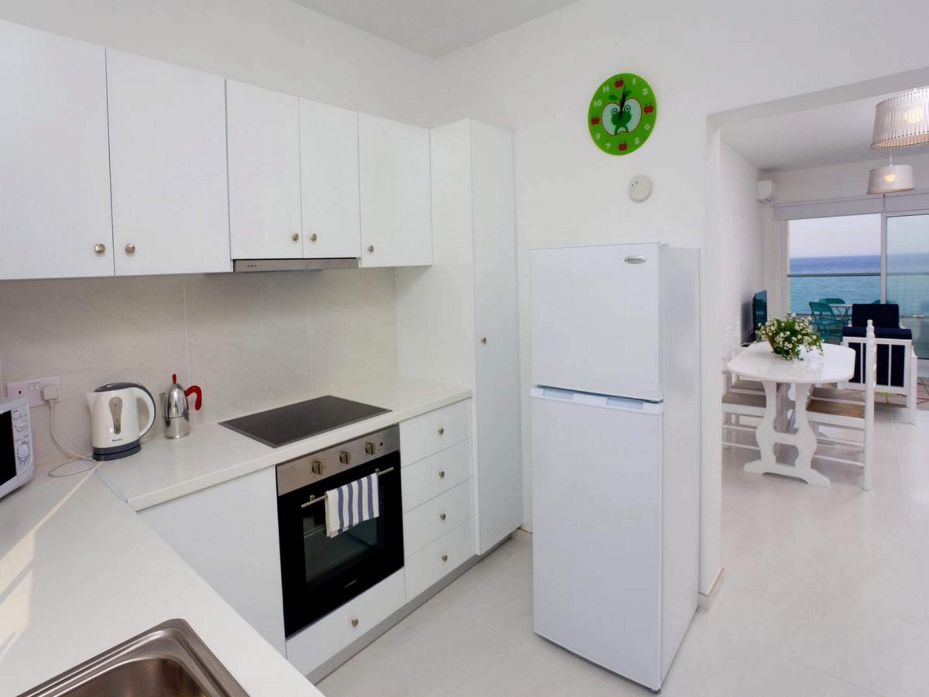 Una cocina moderna con armarios blancos, horno empotrado, refrigerador y vista al mar a través de amplias ventanas.