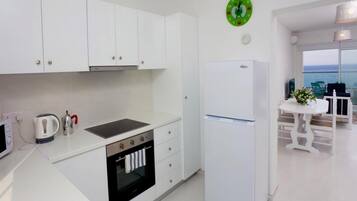 Una cocina moderna con armarios blancos, horno empotrado, refrigerador y vista al mar a través de amplias ventanas.