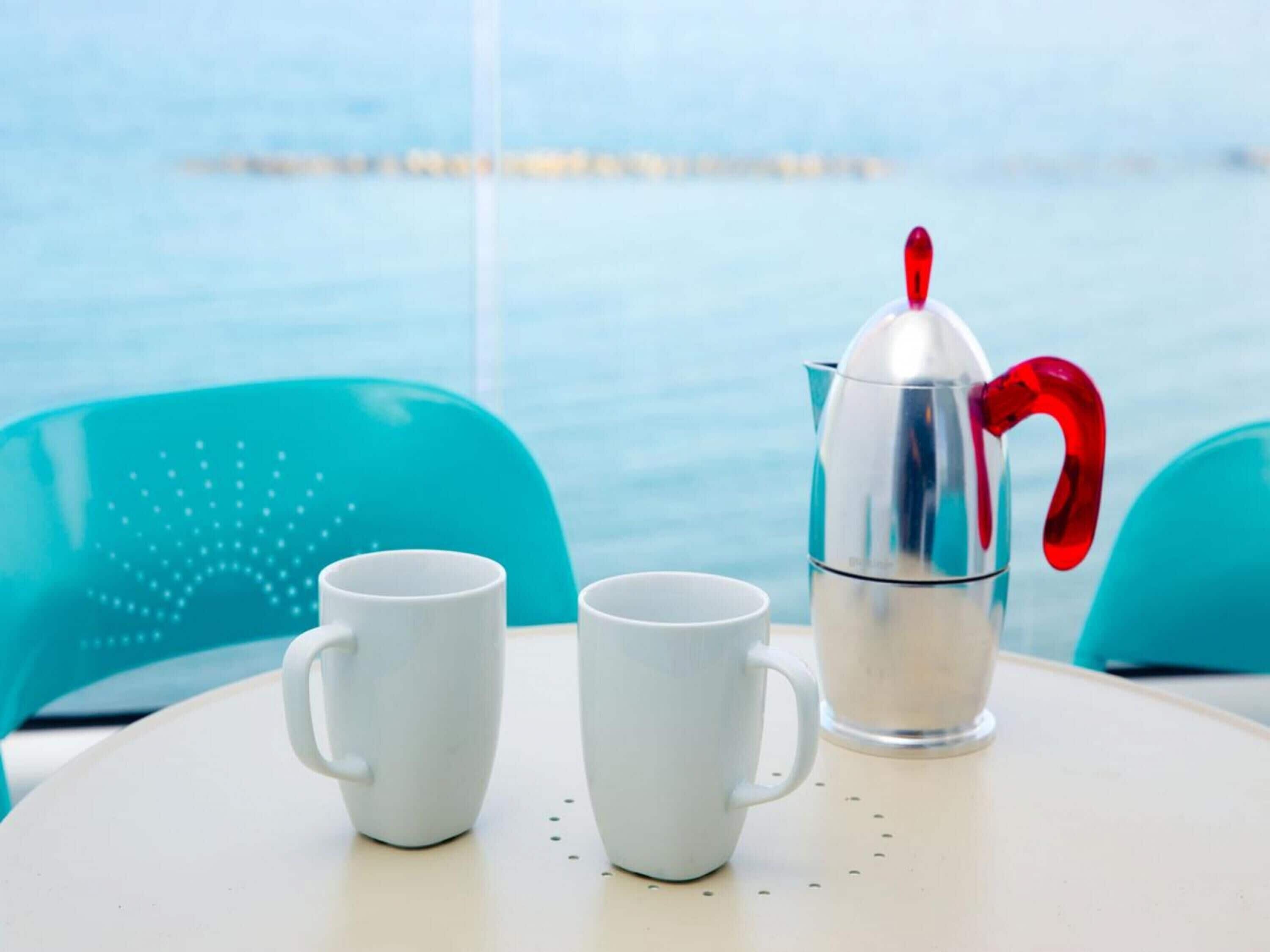 Una mesa con dos tazas blancas y una cafetera plateada con asa roja, frente a una silla azul y una vista al mar.