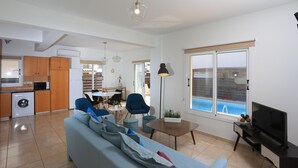 A modern living room with a sofa, a coffee table, and a TV. There is a kitchen area with cabinets and a washing machine. A dining area with chairs and a table is visible. - Seabreeze Villa No 4 (Sotira)