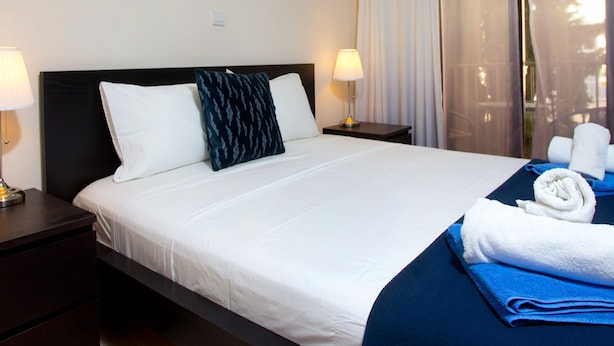 Een hotelkamer met een groot bed, twee bedlampen, een donkere hoofdeinde, een raam met dunne gordijnen en uitzicht op het omliggende landschap.