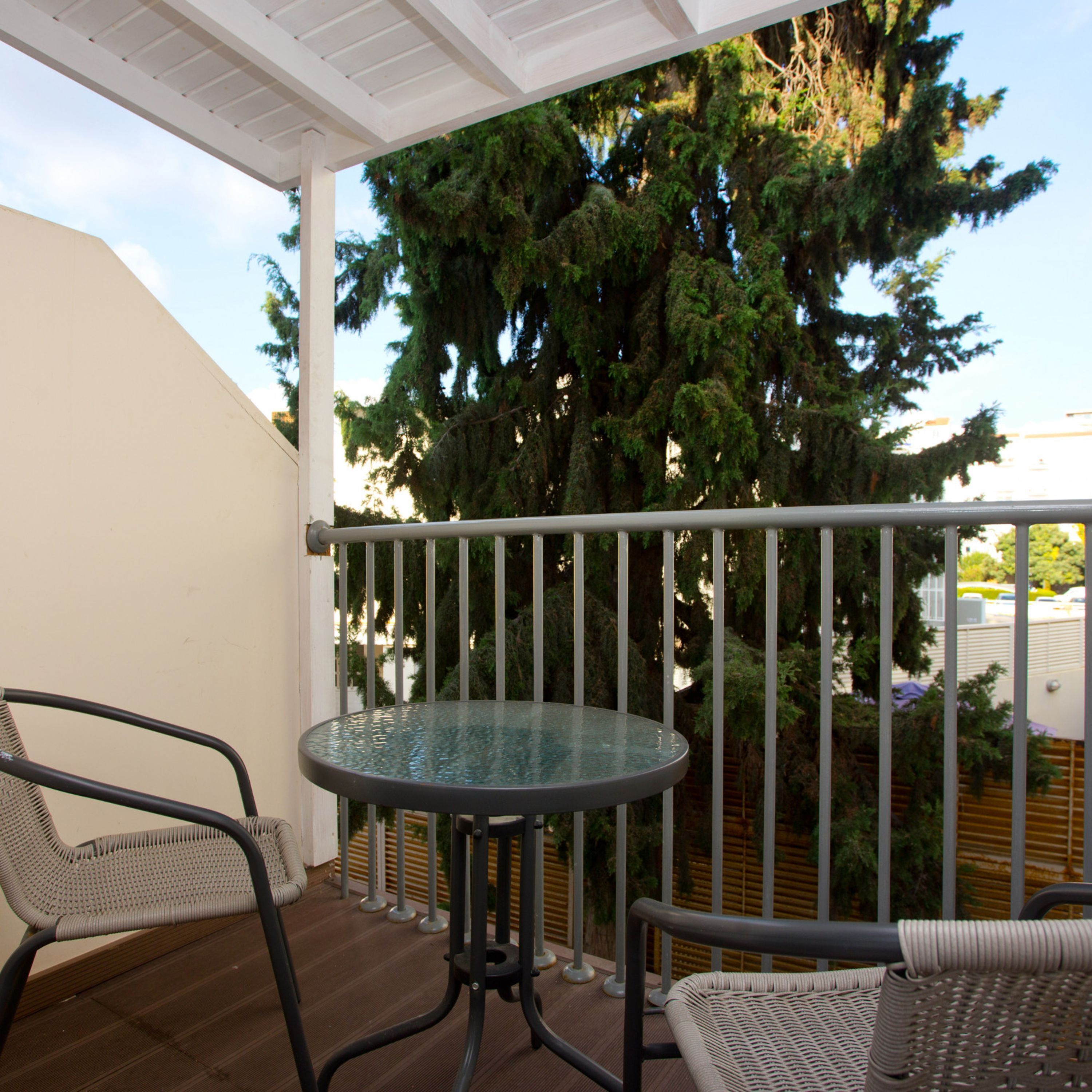 A balcony with a glass table and two wicker chairs, overlooking a large tree and a building.