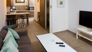 Een moderne woonkamer met een bank, een salontafel en een televisie. Er is een keukenhoek met een eettafel en barstoelen.