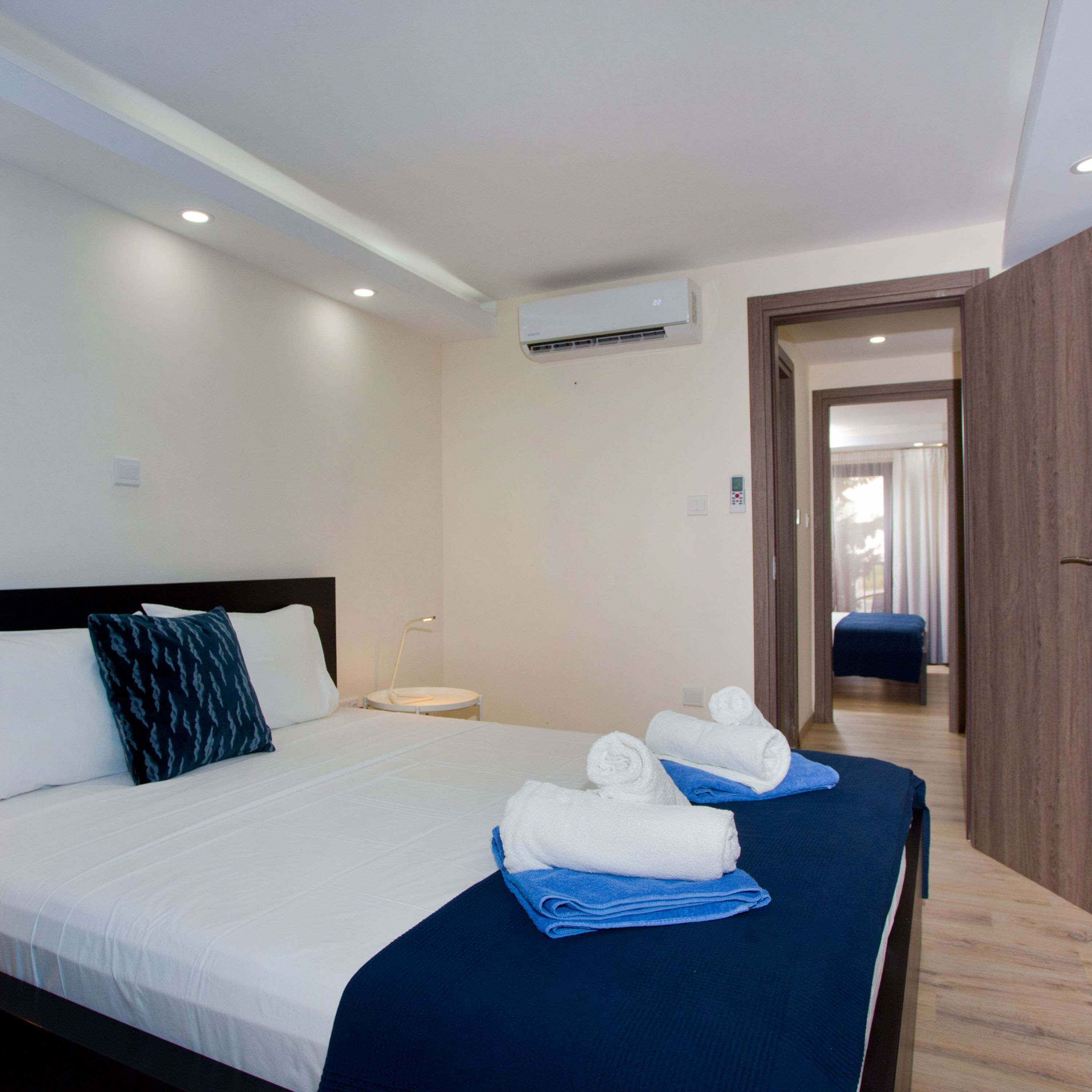 Habitación de hotel con cama, mesita de noche, equipo de aire acondicionado y vista a otra habitación por una puerta abierta.