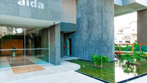 A modern building with a large central column, a glass entrance, and a small water feature with plants. - Opera Exclusive Studio (Limassol)