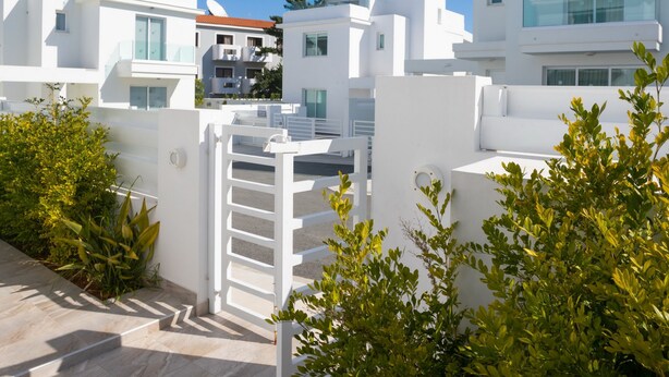 Um edifício moderno e branco, com portão, céu claro e vegetação ao redor.