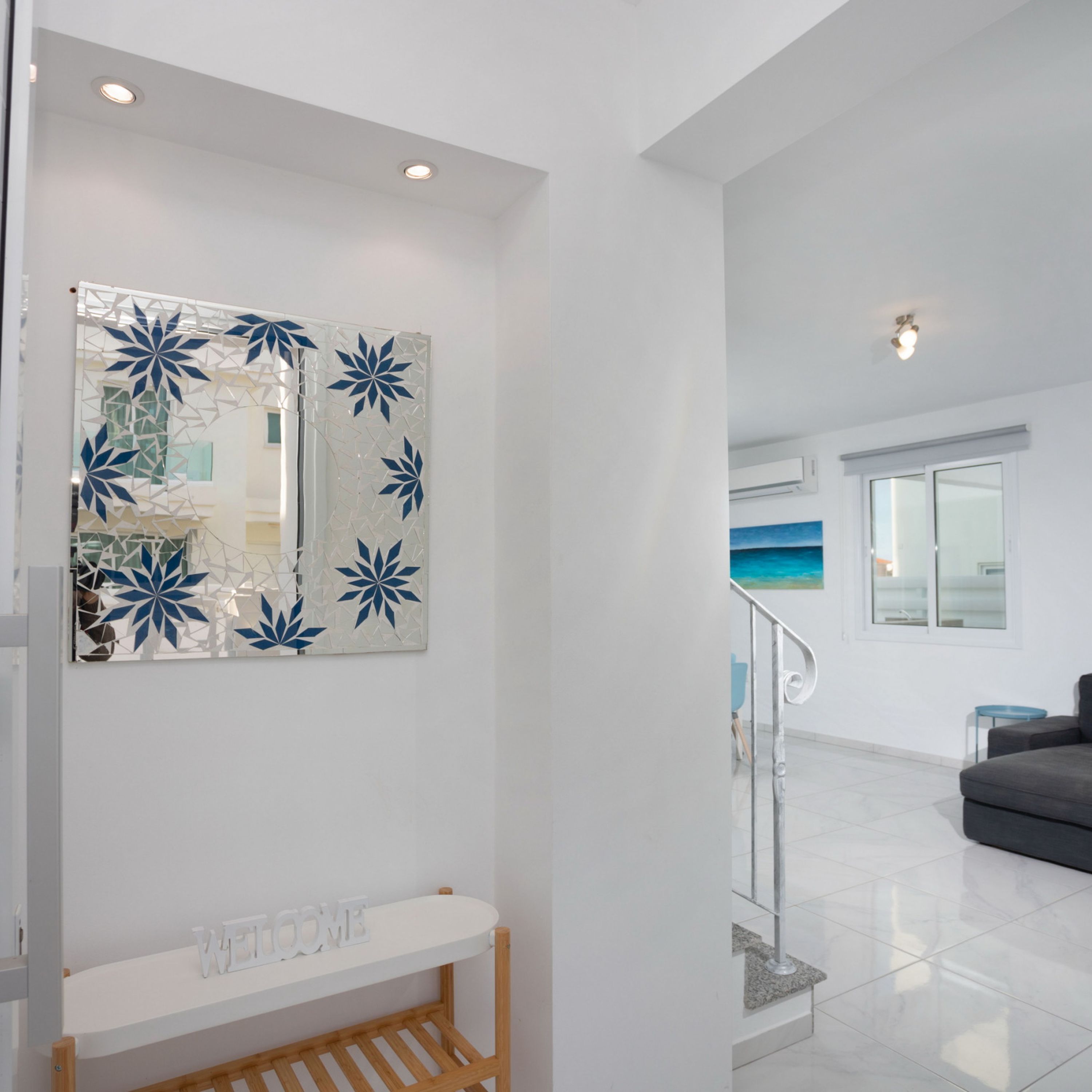 A modern living room with a white wall, a decorative mirror, a white console table, a grey sofa, and a staircase leading to another level.