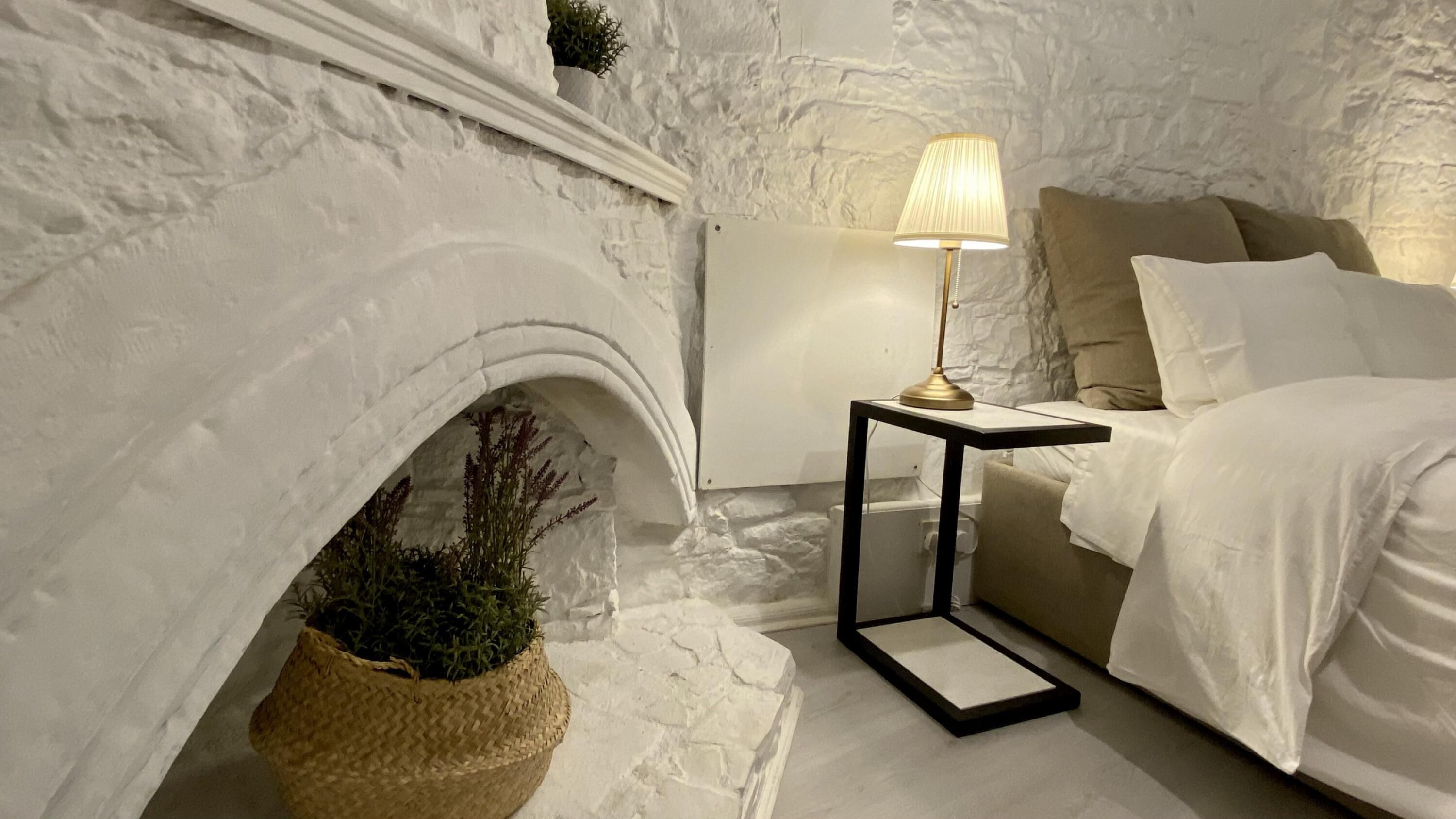 A bedroom with a stone fireplace, a bedside table with a lamp, a bed, and a woven basket with plants.
