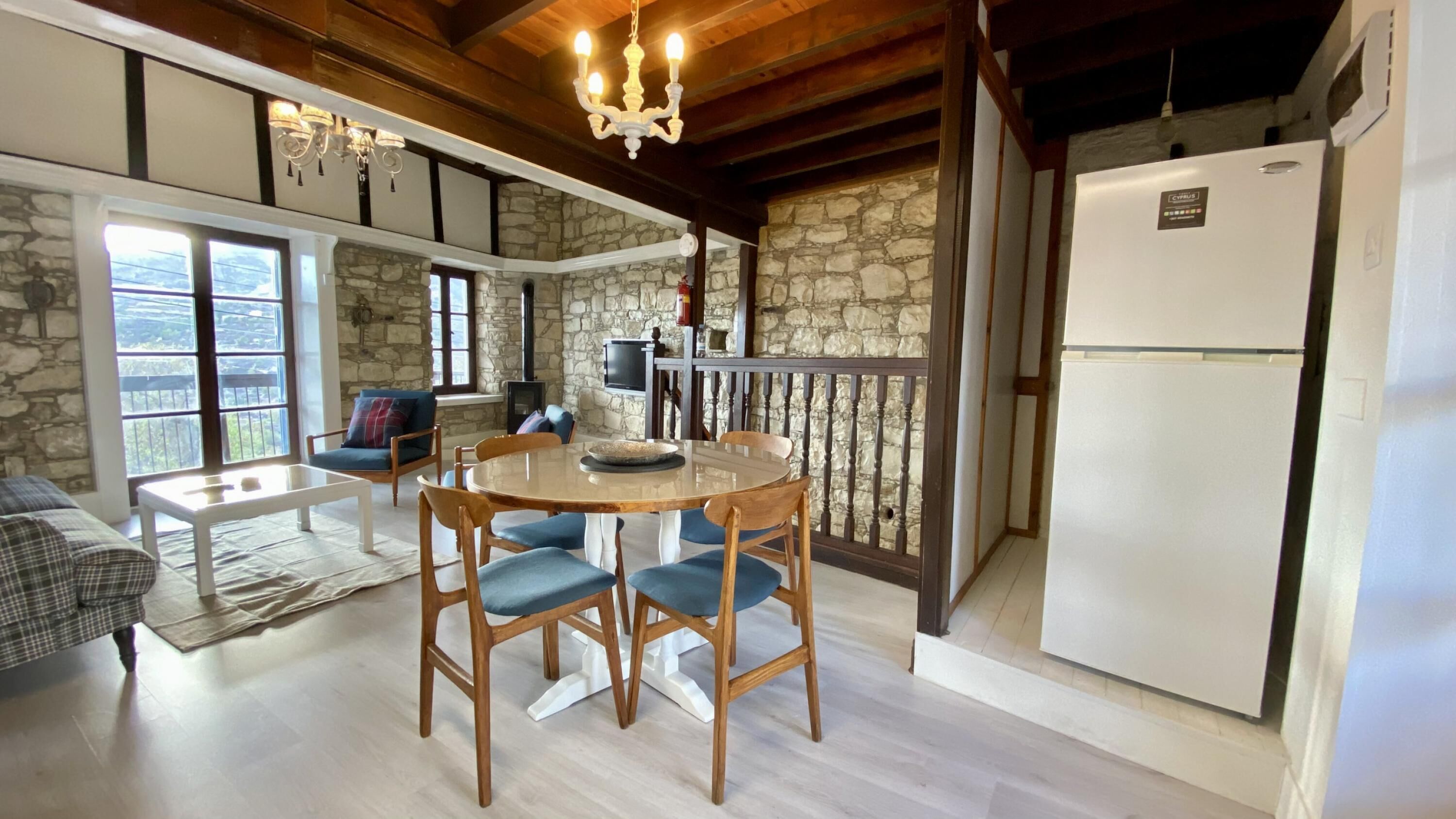 A cozy kitchen with a stone wall, wooden dining table, and chairs. There is a white refrigerator and a TV on the wall.