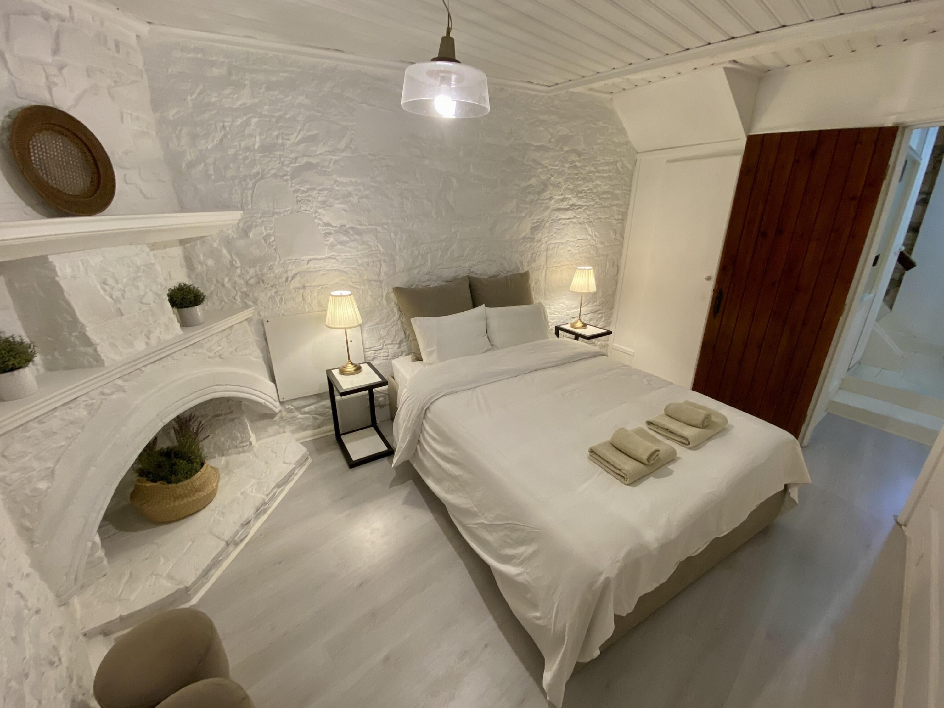 A bedroom with a large bed, two bedside tables with lamps, a fireplace, and a wooden door.