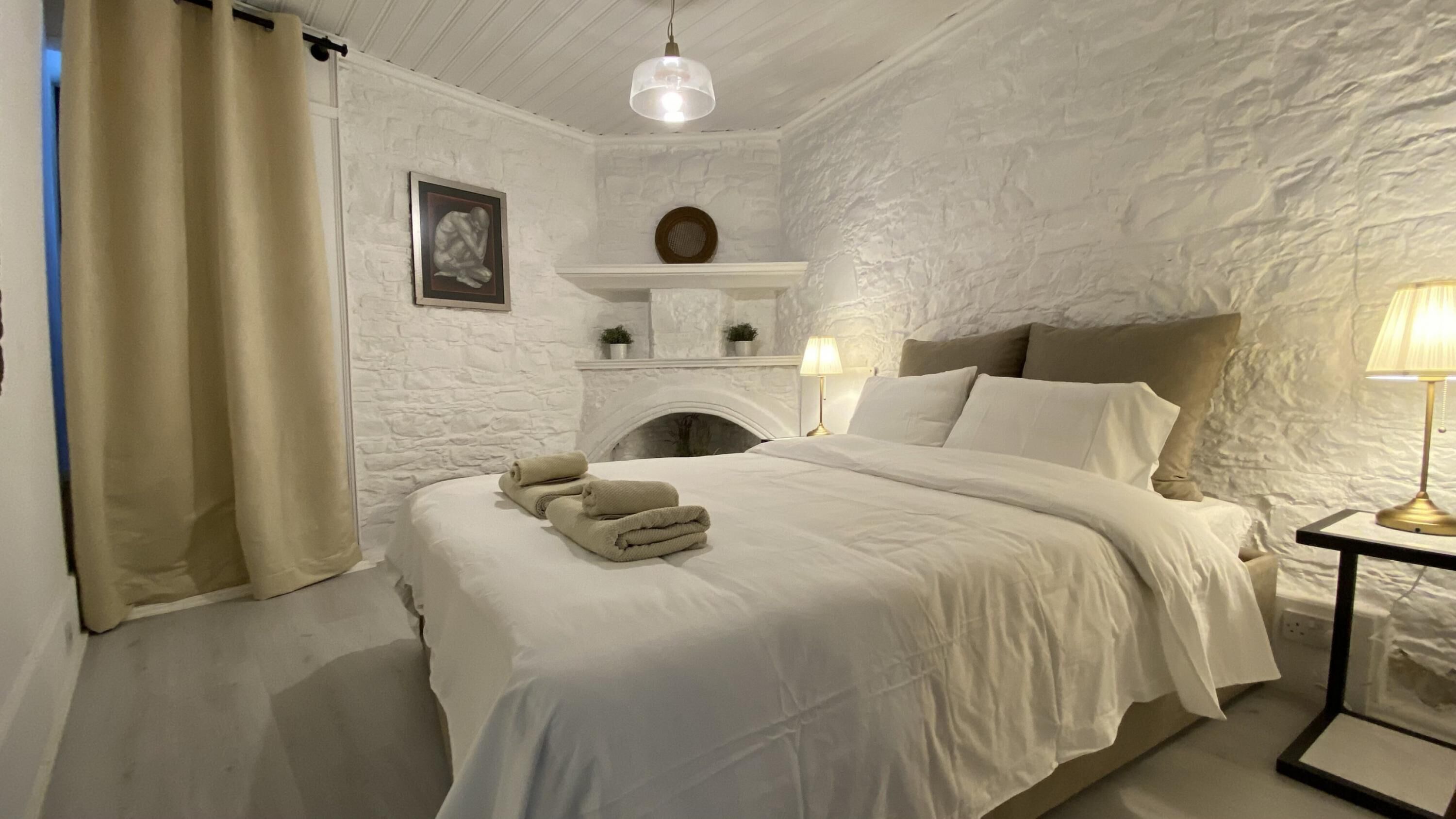 A bedroom with a stone wall, a fireplace, a bed with white linens, and a nightstand with a lamp.