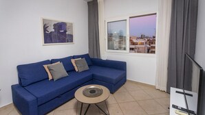 A modern living room with a blue sectional sofa, a small round table, a framed artwork on the wall, and a window with a city view. - Palm Beach Apartment (Limassol)