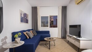 A modern living room with a blue sofa, a small round table, a TV on a white cabinet, and a window with curtains. - Palm Beach Apartment (Limassol)