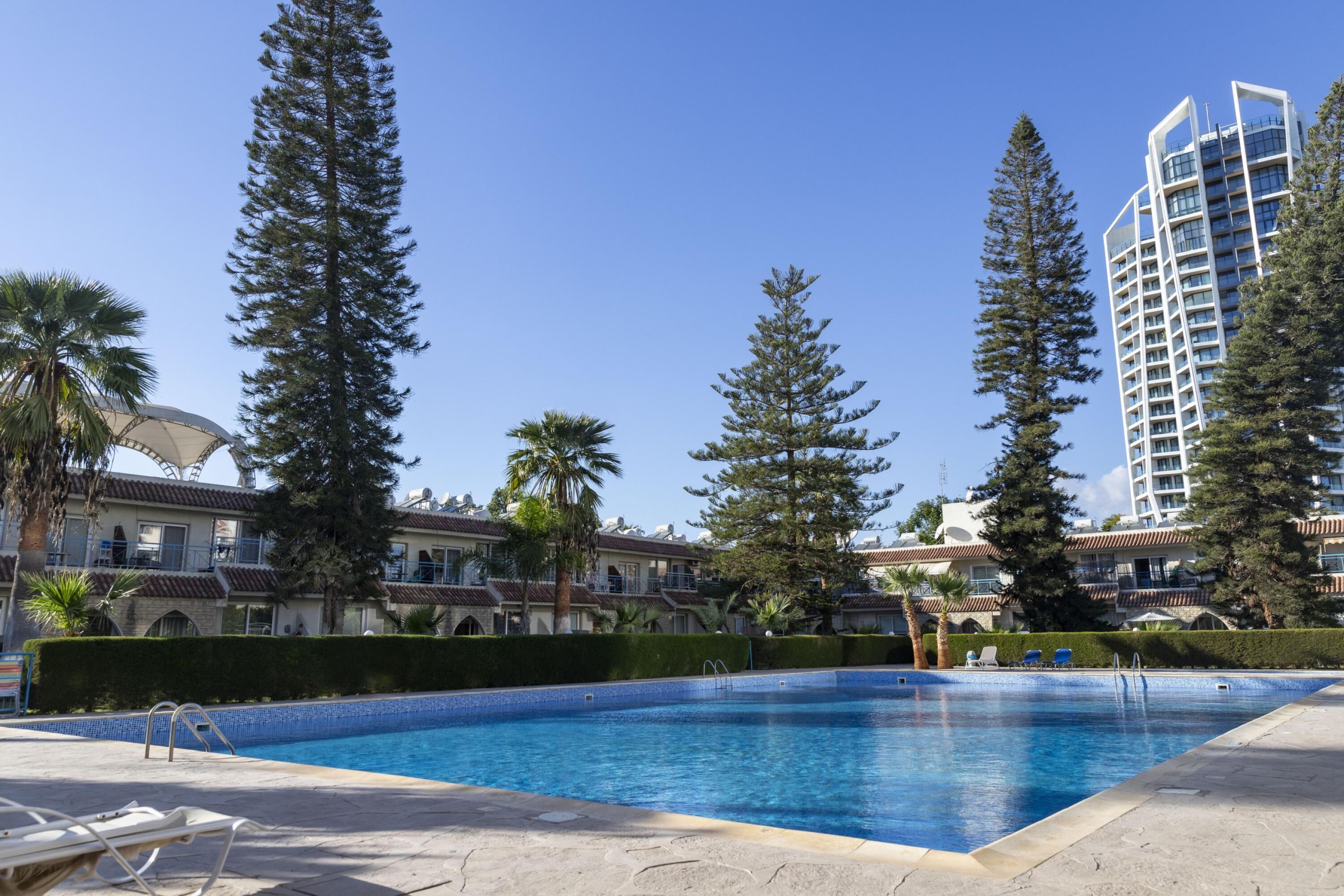 A swimming pool with lounge chairs, surrounded by tall trees and a building with balconies.
