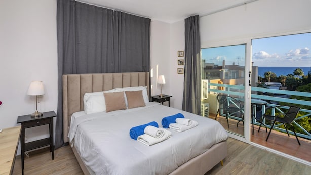 A hotel room with a bed, two nightstands, a lamp, a view of the sea, and a balcony with chairs. - Palm Beach Apartment (Limassol)