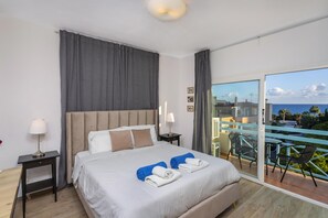 Ein Hotelzimmer mit Bett, zwei Nachttischen, einer Lampe, Meerblick und einem Balkon mit Stühlen.