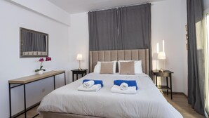 A hotel room with a large bed, two bedside tables with lamps, a bedside mirror, a small desk with a lamp, and a window with curtains. - Palm Beach Apartment (Limassol)