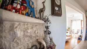A stone fireplace with a shelf holding various decorative items, a wall clock, and a view into a living room with a wooden floor and furniture. - Lania Royal Oak House (Laneia)
