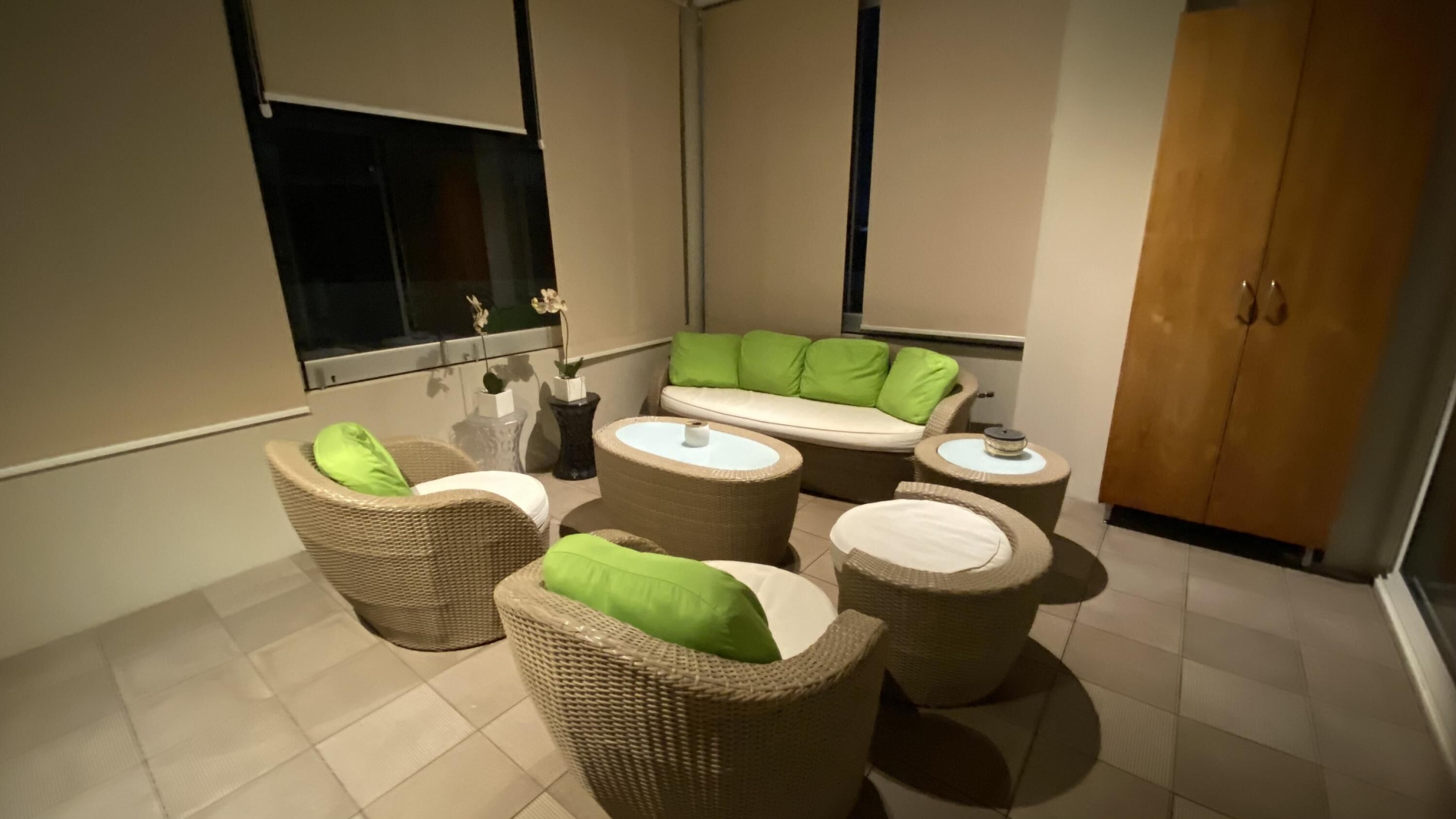 Een moderne badkamer met een zithoek, voorzien van rieten meubels en groene kussens, een grote spiegel en een houten kast.