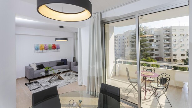 A modern living room with a glass dining table, grey sofa, and a large window with a balcony. - Kanika Seabreeze Apartment (Limassol)