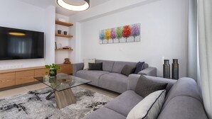 A modern living room with a flat-screen TV, grey sofa, glass coffee table, wooden shelves, and a colorful abstract painting. - Kanika Seabreeze Apartment (Limassol)