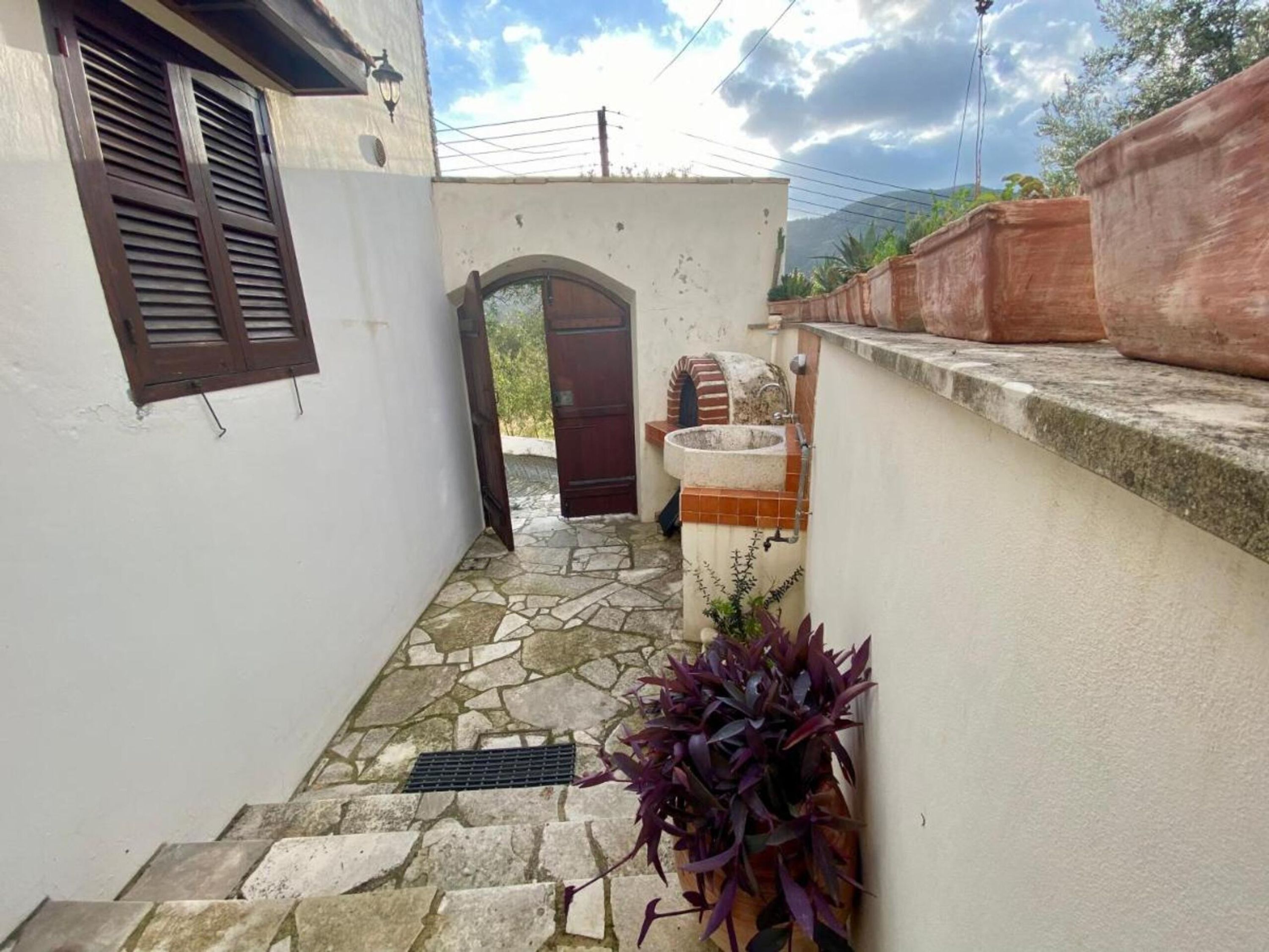 Un estrecho patio empedrado con un muro blanco, una puerta de madera y un pequeño lavabo exterior.