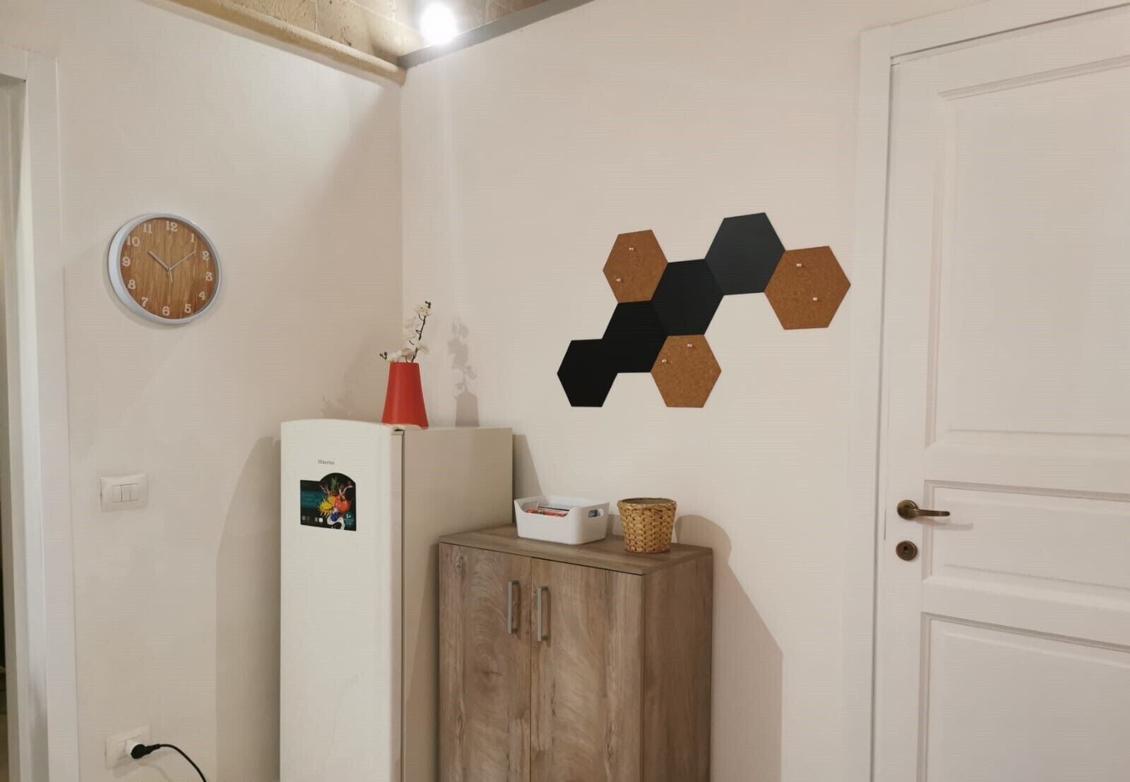 Ein Zimmer mit weißer Tür, einem Holzschrank, einem weißen Kühlschrank, einer Wanduhr und einer sechseckigen Wanddekoration.