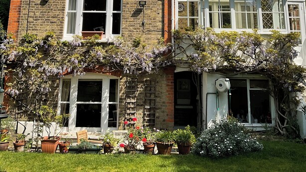 Un edificio de ladrillo de dos pisos con varias ventanas, un porche cubierto y un jardín con plantas en macetas.
