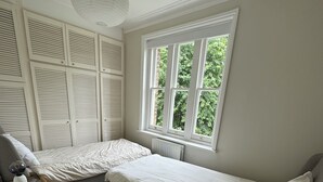 Un dormitorio con dos camas individuales, una mesita de noche de madera con lámpara, un radiador y un ventanal con vistas al follaje.