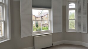 Habitación con dos ventanas, una con contraventanas abiertas y otra cerradas, un radiador debajo de la ventana abierta, y vista a una casa de ladrillo y follaje exterior.